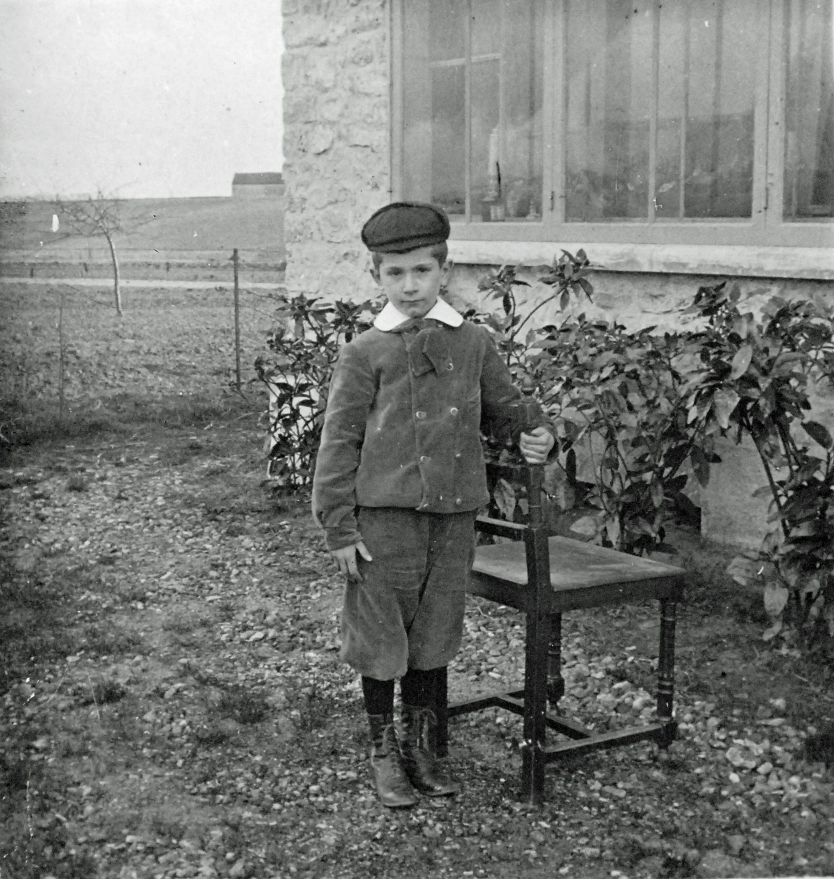 Garçon à casquette appuyé sur une chaise devant une façade en pierre et une baie vitrée