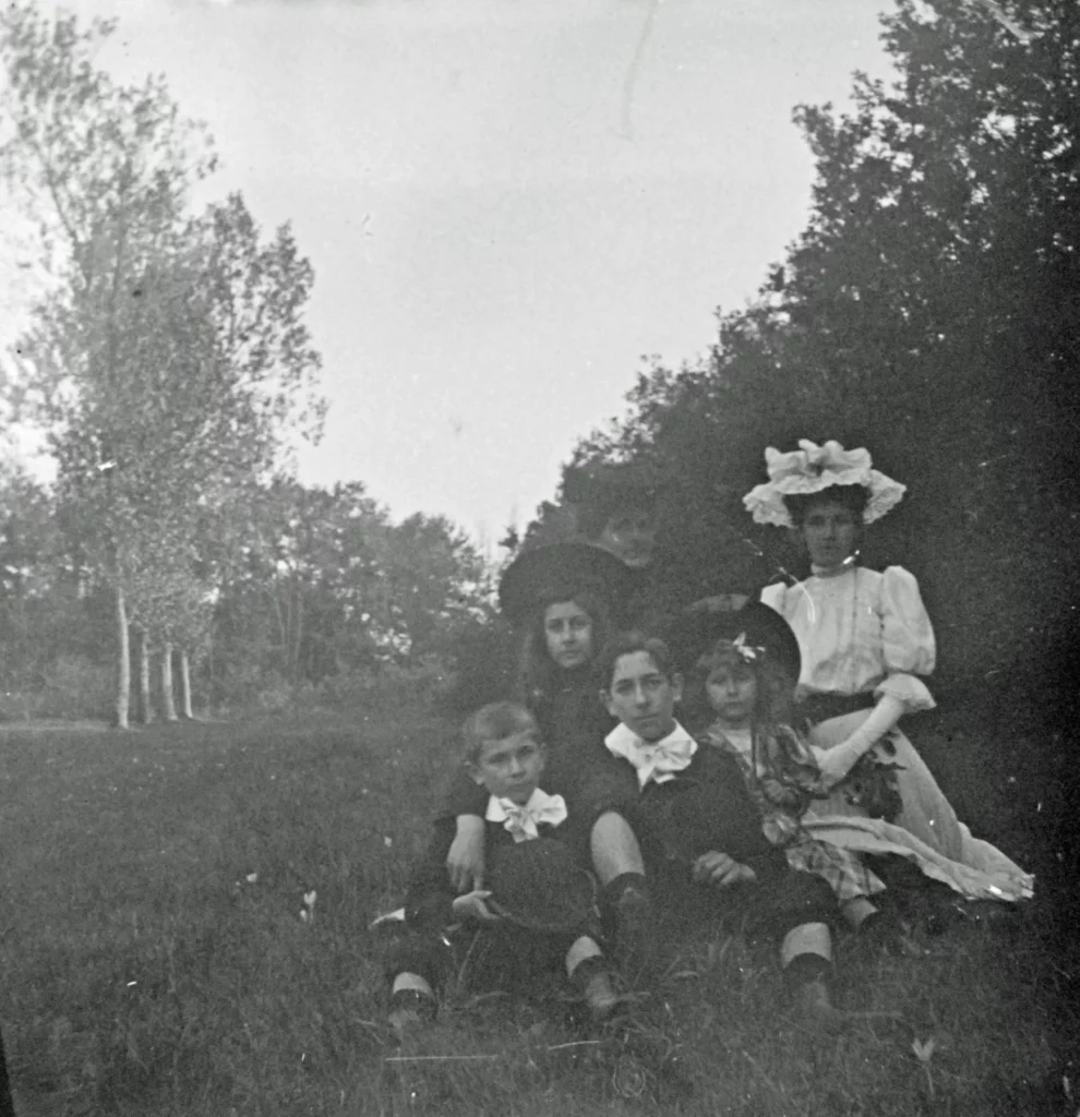 Enfants groupés dans l'herbe autour d'une jeune femme en robe blanche et grand chapeau